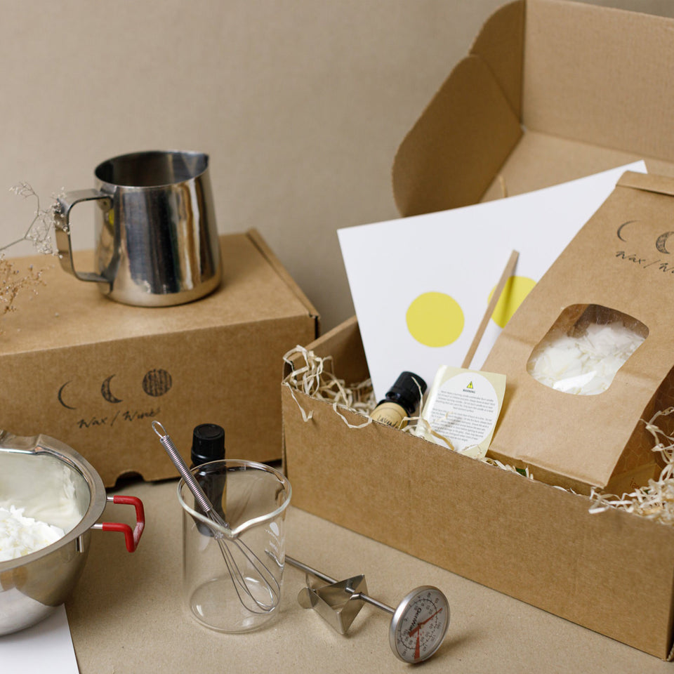 a couple of cardboard boxes containing a bowl with flaked soy wax, a beak glass, a thermometer, a metal jug, a small amber bottle and a whisk